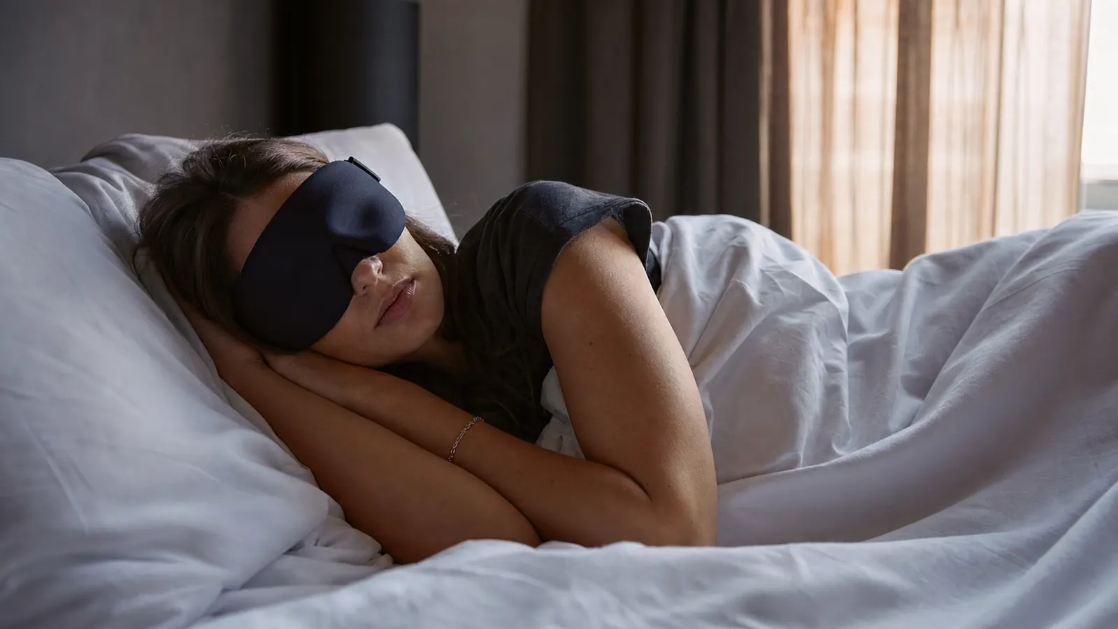 Person sleeping on their side wearing the MZOO 3D eye mask, room in near-total darkness with blackout curtains partially drawn