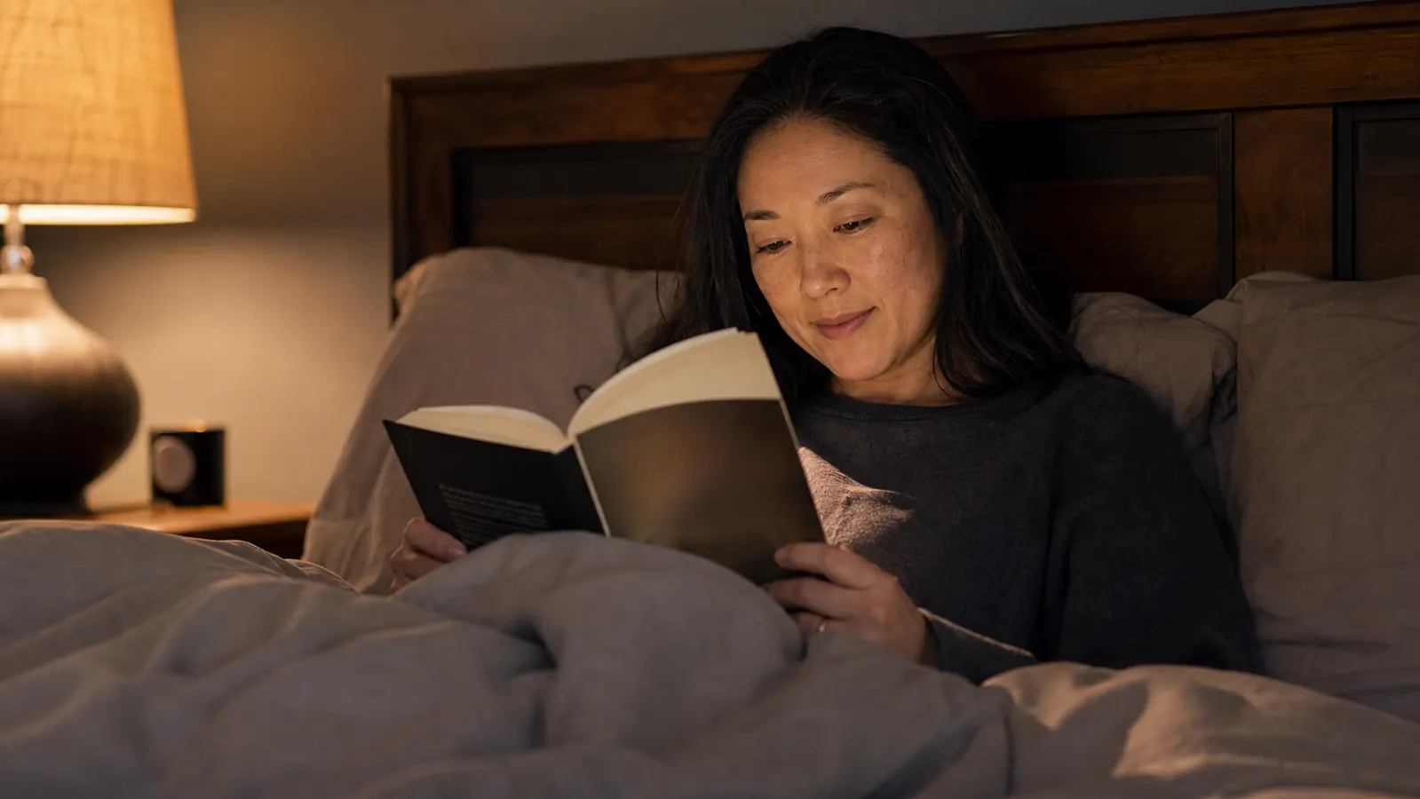 Person reading quietly in bed under a weighted blanket, lamp on the nightstand casting a warm glow